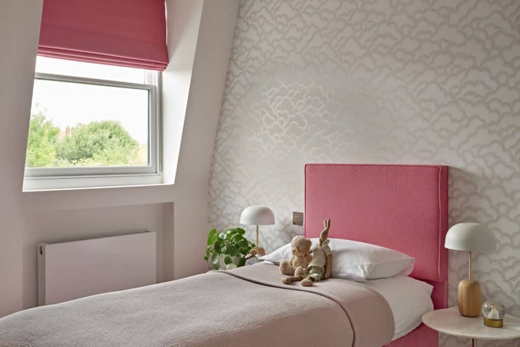 A luxury cute and cosy pink bedroom. The walls adorned with white cloud patterns creating a dreamy atmosphere. A single bed with a pink bed base and matching headboard takes centre stage, dressed in white bedsheets and adorned with a pink throw and two adorable teddy bears. Marble bedside tables either side of the bed, accompanied by white domed lights with wooden bases. The tables are adorned with plants and accessories. Positioned next to a window, the matching blind creates a cohesive look with the headboard.