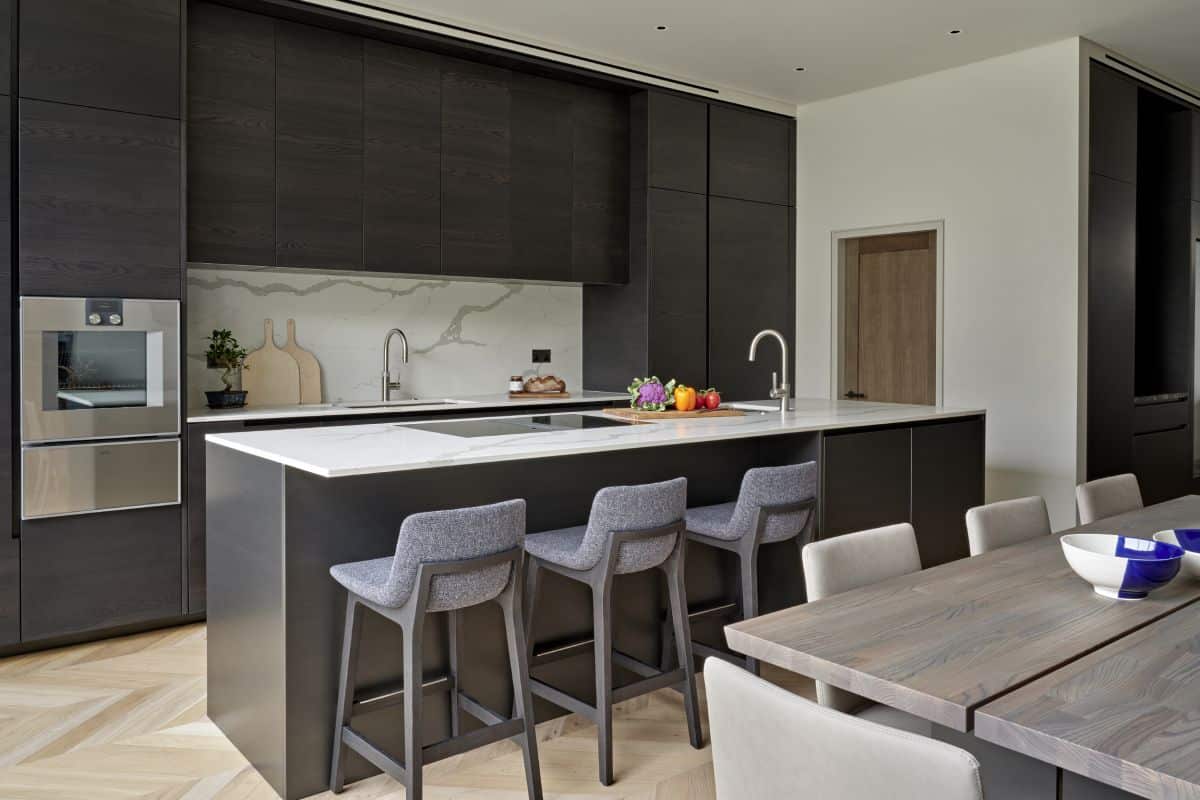 A luxurious modern, open plan kitchen, dining room designed to impress. A white marble top island takes canter stage, featuring a sleek cooker and a sink. The white marble back countertop showcases stylish kitchen accessories, adding both functionality and elegance. The dark wood joinery, matching the base of the island, brings a touch of richness to the space. Three grey counter chairs line up next to the island, offering a cosy spot for casual dining. Light wood herringbone flooring adds warmth and texture to the room. A light wood dining table, accompanied by white leather dining chairs, creates a sophisticated setting for formal meals. This luxury kitchen is a perfect blend of style, functionality, and comfort.