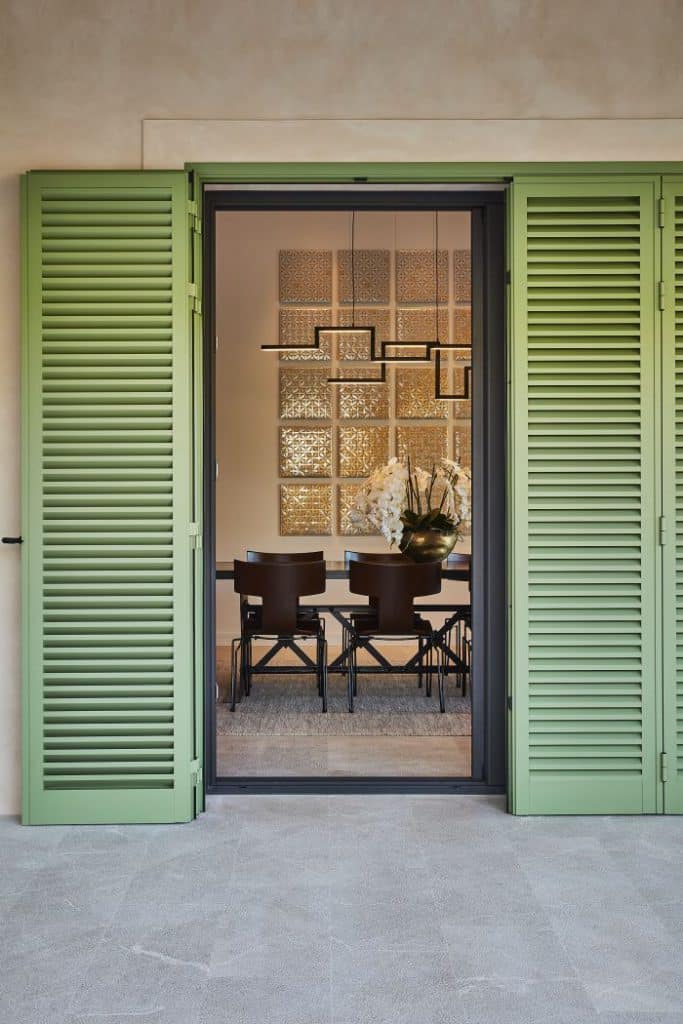 Open sage green shutters on Mallorca villa offer a view into dining room, where a set of Rubelli leather anziano dining chairs are sitting around a dining table.