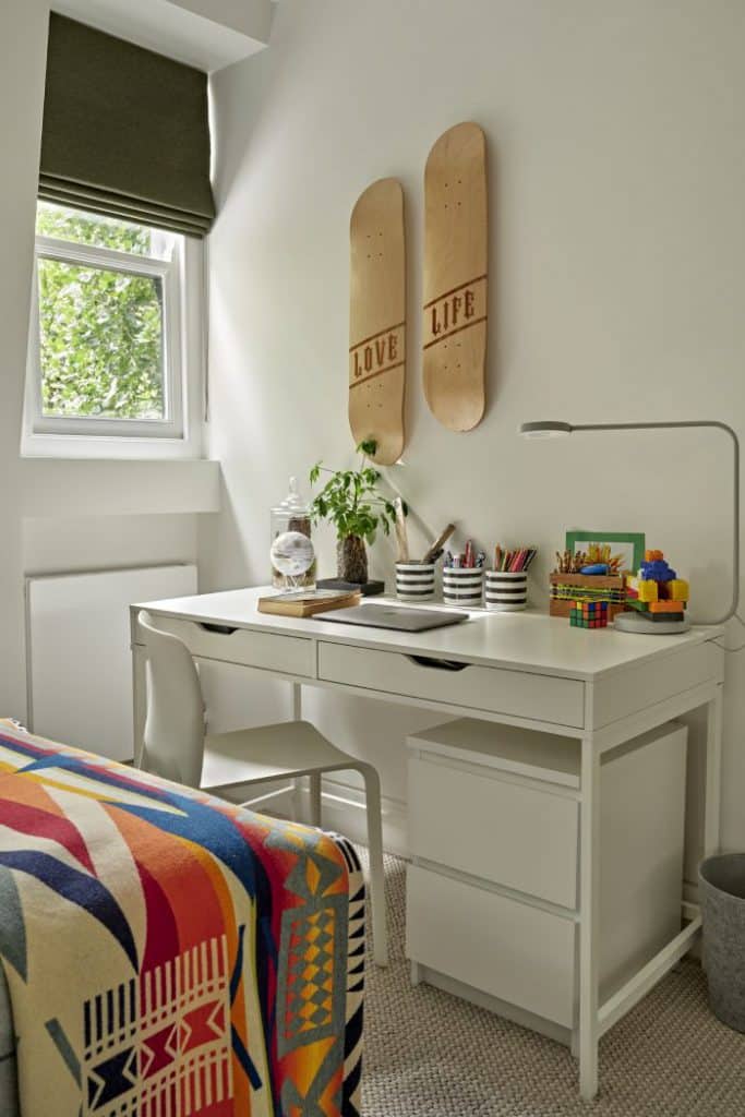 A luxurious boys' bedroom with a study area. A sleek white desk with storage space is accompanied by a matching chair, offering a comfortable and stylish workspace. The desk is adorned with essential items such as a book, laptop, writing equipment, a vibrant plant, and a desk light. Above the desk, two skateboard bases hang proudly on the pristine white walls. Positioned next to a window, adorned with a green blind, natural light fills the space. The end of the bed is showcased, presented with a colourful patterned blanket adding a hint of character.