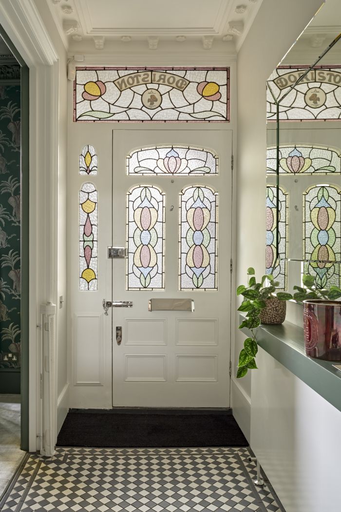 A luxurious front door and hall, greeted by the beauty of a white stained glass Victorian home door, exuding elegance. A black doormat lies gracefully at the entrance. The floor is a stunning white and black chequered tile design, capturing a sense of classic style. The walls, painted in pristine white, create a bright and welcoming ambiance, complemented by a large mirror mounted on one wall. Below the mirror, a grey shelf adorned with lush green plants adds a natural element.