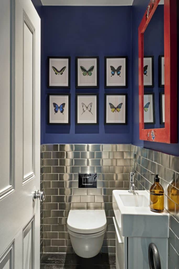 This luxurious bathroom. The bottom half of the wall is adorned with sleek stainless steel tiles. The top half is painted in a deep, elegant shade of dark blue, creating a dramatic contrast. A white toilet with a matching silver flush is accompanied by a white sink featuring a sleek silver tap. Above the toilet, six individually framed images of butterflies. Completing the design, a mirror with a bold red frame hangs above the sink.