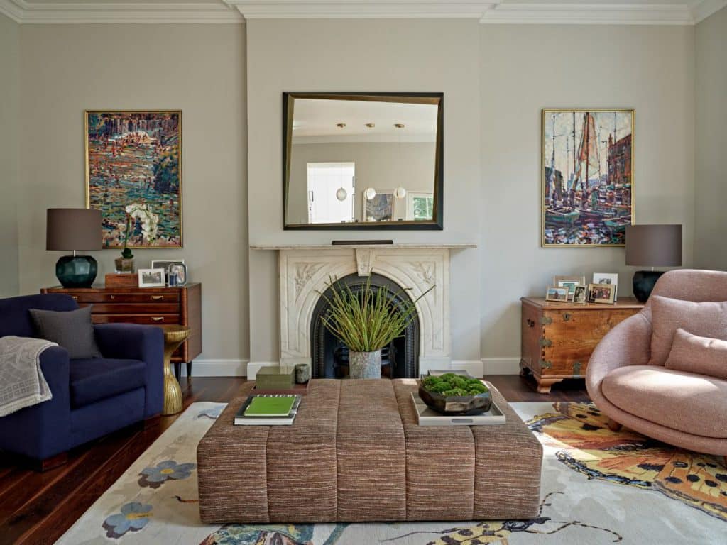 A luxurious lounge filled with character. The dark wood floor complemented by a cream painted ceiling overhead. A rug adorned with intricate patterns adds a touch of sophistication. One wall a magnificent Tom Faulkner mirror above a white period fireplace, with a vibrant green plant positioned in front, adding a natural element. In the centre of the room, an ottoman crafted from brown textured fabric invites relaxation, adorned with a stack of books. In one corner, a light burgundy armchair with a blue throw sits next to an old wooden chest with picture frames and a lamp adding a touch of personality to the room, with a framed painted image above. In the opposite corner, a blue armchair with a cosy grey throw is accompanied by a brown chest of drawers, housing another colourful framed painting.