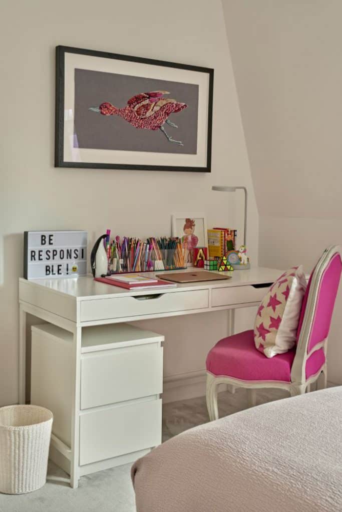 A luxury pretty pink girls bedroom study area. A white desk takes centre stage, adorned with a delightful collection of pens and books, accompanied by an elegant vibrant pink chair with white legs, adding a pop of colour, adorned with a white and pink stary cushion. Above the desk, a framed image of a pink bird, housed in a sleek black frame, captures attention with its graceful beauty.