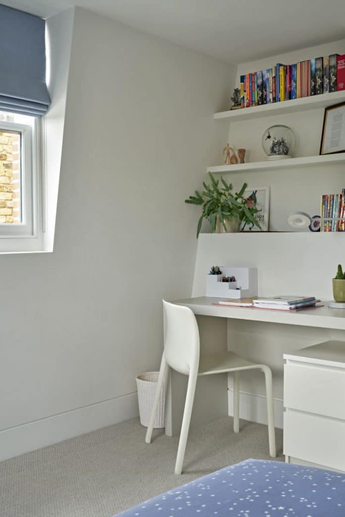 Luxury Children’s bedroom with a cosy study area. A pristine white desk adorned with writing equipment and books takes centre stage, accompanied by a matching white chair. Above the desk, elegant white shelves display a collection of books, plants, and personal items, adding a touch of personalization to the space. Positioned next to a window, the study area is surrounded by natural light.. The light blue accents in the window blinds and bed sheets provide a refreshing ambiance.