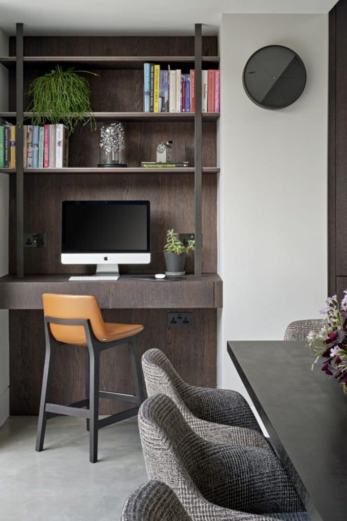 A luxurious open plan study area which leads into the kitchen. The grey stone floor sets the stage, while the brown wood patterned wallpaper divides the study area in. A built in desk with matching wood finish accommodates an Apple computer, accompanied by a plant. Above the desk, three shelves showcase a selection of books, plants, and accessories. A brown leather chair with dark wood legs adds sophistication. The remaining white walls create a sense of serenity, while hanging a black clock. Nearby, a dark wood kitchen island adorned with grey patterned stools and delicate flowers.