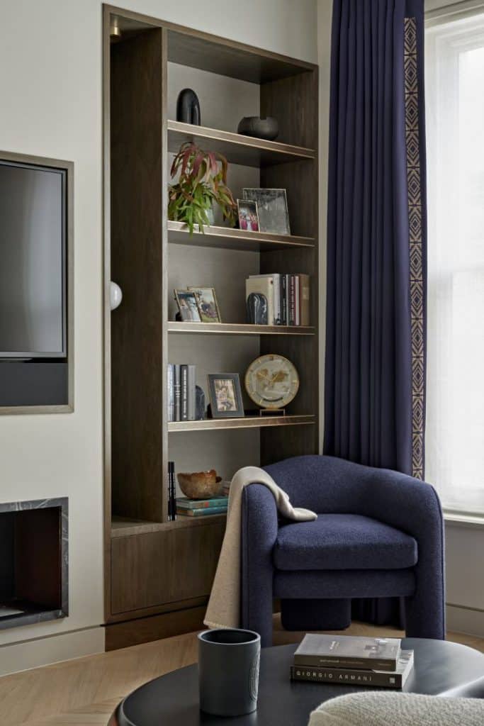 This modern luxury living room features lightwood herringbone flooring. A wall-mounted TV sits above a sleek black marble-framed fireplace. On the right-hand side of the TV, dark wood shelving, by Lethbridge bespoke joinery, built into the wall, adorned with books. In the centre of the room, Ben Whistler bespoke, purple armchair with a cream throw draped over one arm, accompanied by a black circular coffee table displaying books. Adjacent to a large window dressed in white blinds and purple curtains.