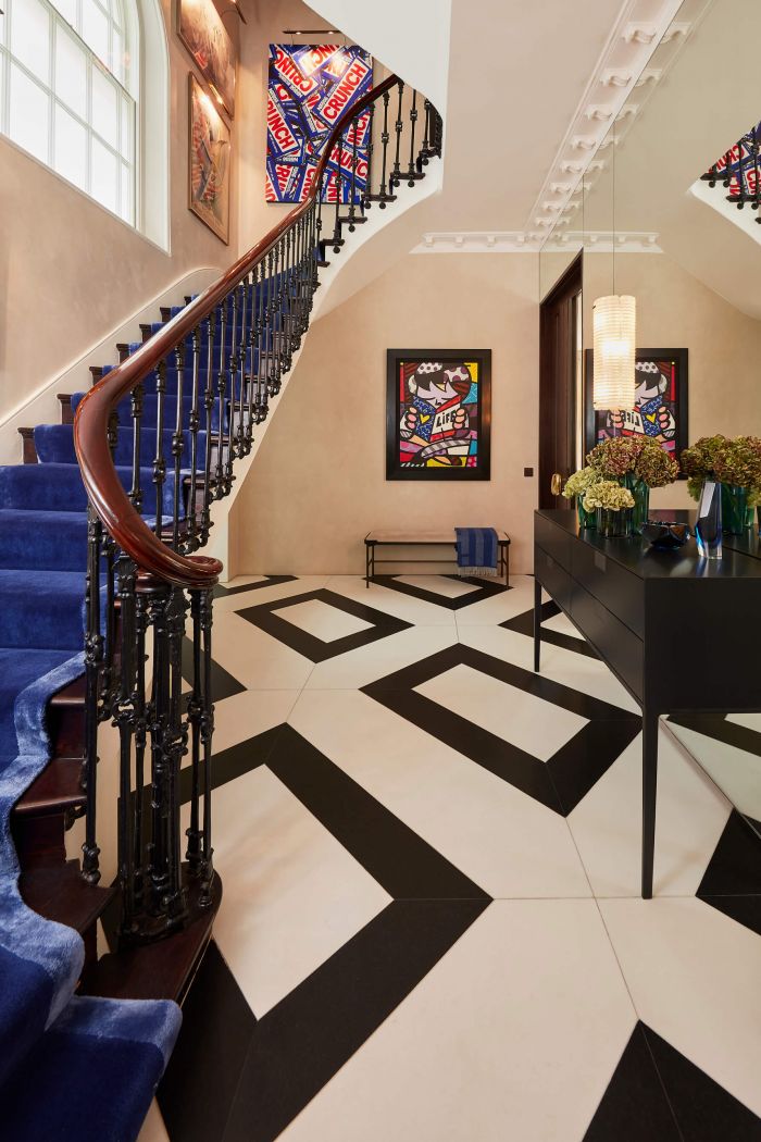 A luxurious striking hallway. The Venetian plaster walls exude elegance, complemented by a black and white diamond-shaped tiled floor. Hanging on the far wall is a vibrant painting in a sleek black frame, accompanied by an ottoman with black legs and a plush blue throw. On the right, a mirrored wall reflects the beauty of the space, featuring a doorway and a stylish black console with pull out drawers, adorned with a collection of flowers. Suspended above is a pendant light. Opposite the mirror wall, a grand period staircase by Henry Van Vijiver, with royal blue carpeting running up it, while large windows bathe the stairs in natural light. At the top of the staircase, a collection of abstract art pieces illuminated by projected lights.