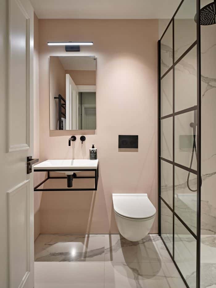 A luxurious bathroom adorned with baby pink walls and a pristine white marble tiled floor. With a white toilet complemented by sleek black fixtures. To the left, a white elegant sink with black fixtures and a structured design offers a sophisticated touch. Above the sink, a rectangular mirror is illuminated by a bright light, enhancing the space. In the opposite corner, the shower showcases exquisite marble tiles that match the floor, complete with black fixtures and a glass screen framed in black.