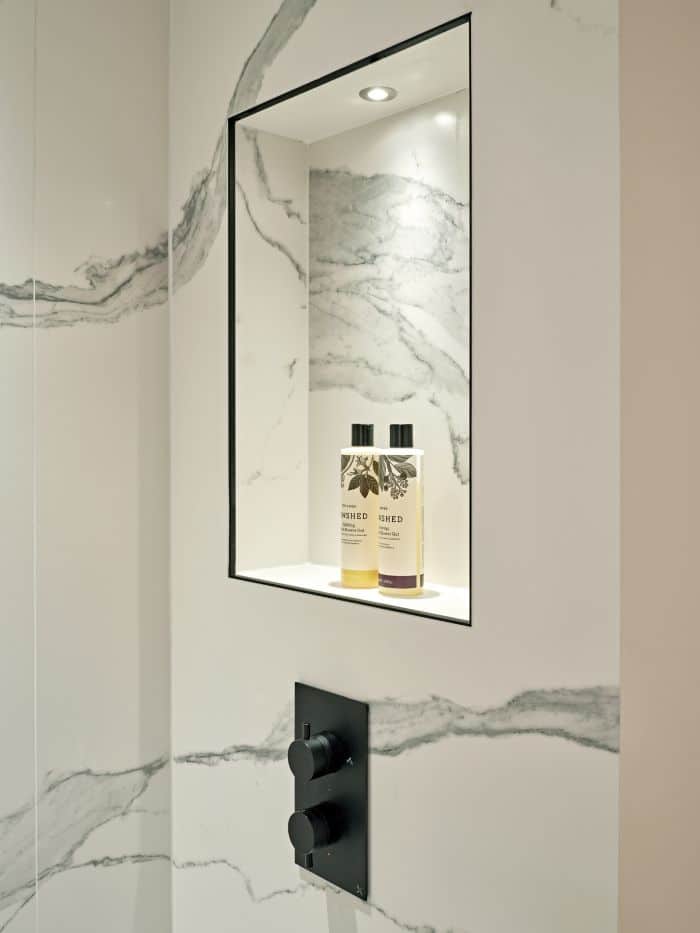 A close up image within a luxury bathroom. White marble tiles with grey veins running through them , adorn the walls creating a brilliant bright tranquil aesthetic. The focal point of this image is the built in shelf also made from marble, this shelf is framed in black and has a convenient light, adding a touch of functionality, displayed on the shelf tasteful bathroom accessories. Below the shelf complementing the framing, black fixtures are on display, creating a sense of uniform in the space.