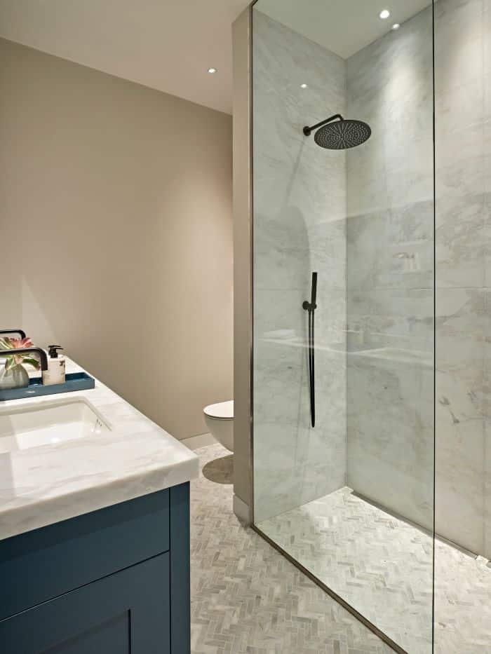 A luxurious, exquisite bathroom. Crisp white walls provide a pristine canvas, while a grayscale herringbone marble tiled floor adds a touch of elegance. In one corner, a dividing wall conceals the toilet, offering privacy and functionality. On the opposite side of the wall, a white marble shower adorned with sleek black hardware and a glass screen. Positioned across from the shower, a his and hers sink with a white marble countertop with a stylish blue painted base, adorned with black taps and tasteful bathroom accessories.