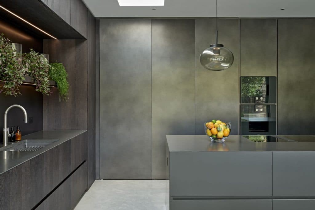 This luxury kitchen features light grey stone-look flooring, providing a sleek and sophisticated foundation. Dark wood kitchen cabinets and joinery, paired with a slate black countertop and sink, create a striking contrast. Above the sink, shelves adorned with lush plants add a touch of freshness to the space. On the opposite wall, slate black cabinets and a well-organised pantry door enhance functionality and organisation. A central slate black island houses hobs and is topped with a large glass bowl filled with vibrant oranges, lemons, and limes. Above the island, a grand glass bulb light serves as a captivating focal point.