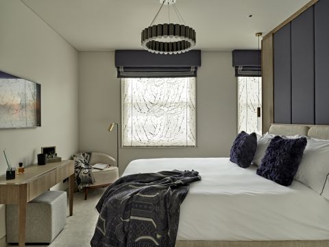 A luxury master bedroom. A wooden room divider doubles as a headboard, adorned with panels of grey and purple fabric. The bed boasts a secondary headboard in a soothing beige tone, complemented by white bedding adorned with two elegant purple accent cushions and a patterned throw. At the foot of the bed, a wooden table with a cosy grey ottoman, while above, a wall-mounted TV. A cosy armchair in the corner, accompanied by a soft throw and a floor lamp for reading. The windows have purple blinds matching a magnificent circular chandelier above the bed.